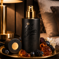 A matte black perfume bottle with a gold spray top sits on a gold tray, surrounded by amber and dark berries, with a matching cap placed beside it. The setting includes a warm lit lamp, dark wood background, and pillows, creating a luxurious bedroom-like environment.